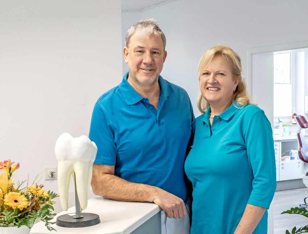 Zahnärztin und Zahnarzt in türkisfarbener Kleidung am Empfangstresen mit Zahnmodell und Blumendeko.